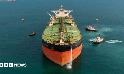 Stock image of an oil tanker pictured front on, with several tugs surrounding the ship
