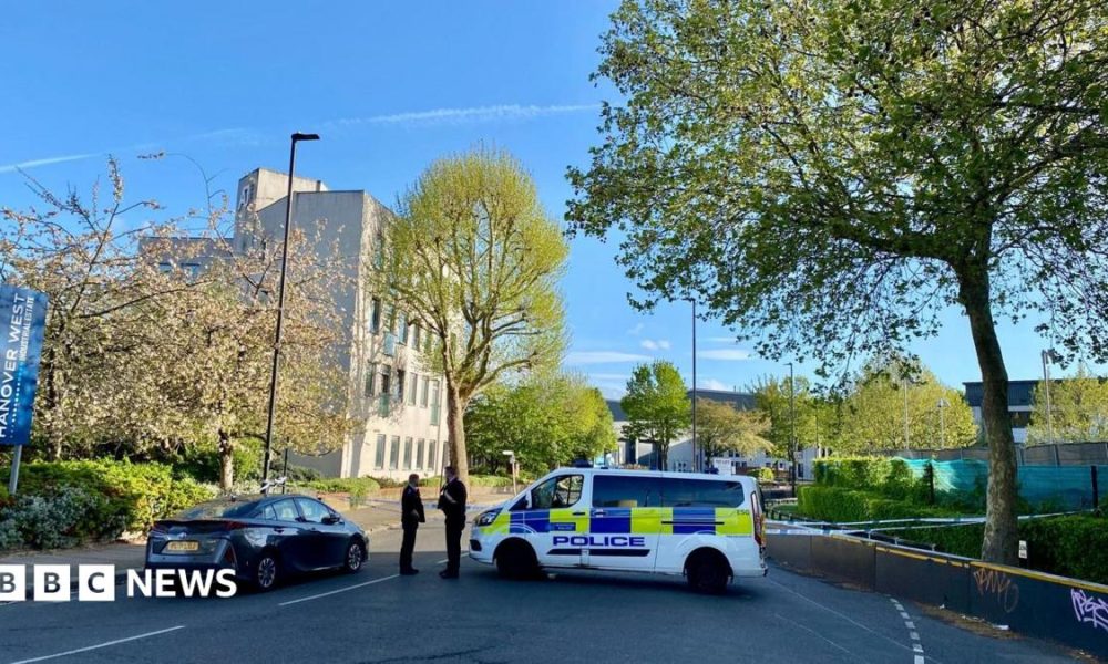 Police tape closes off the road. Two officers can be seen standing next to a police vehicle. The area is leafy with a pale building in the background.