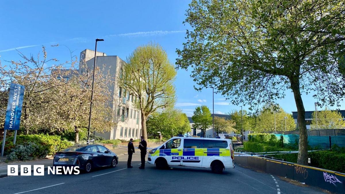 Police tape closes off the road. Two officers can be seen standing next to a police vehicle. The area is leafy with a pale building in the background.