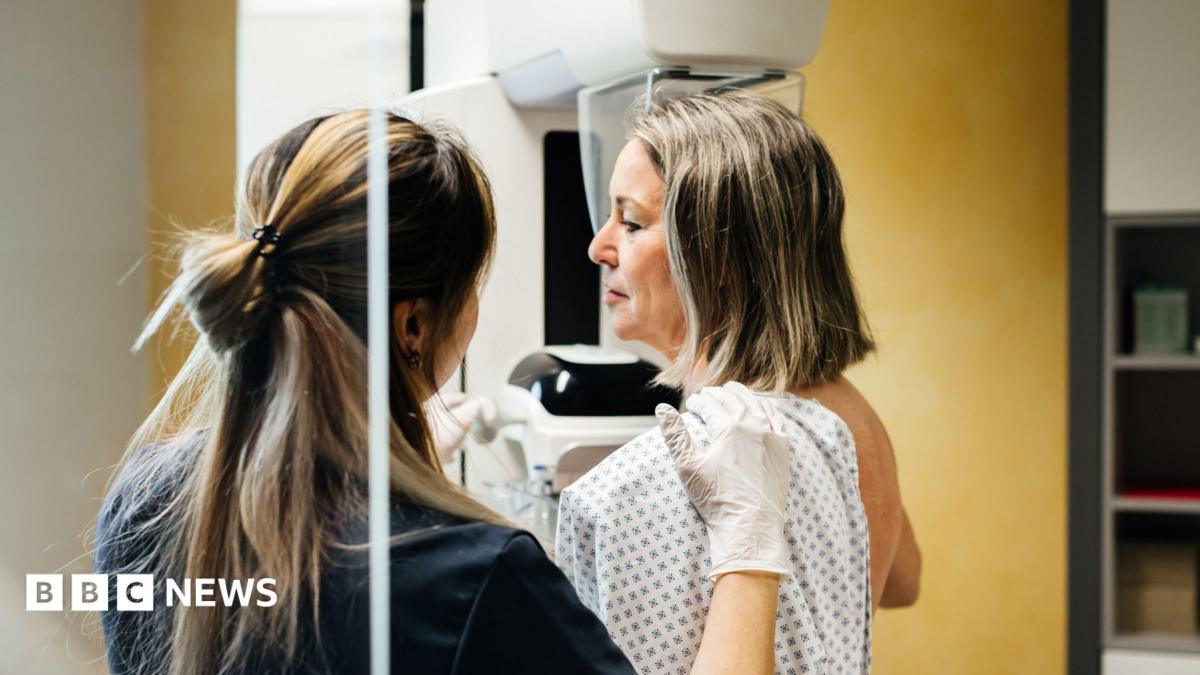 Healthcare professional wearing white examination gloves positioning a patient in a medical gown for a breast imaging scan using mammography equipment in a clinical setting.