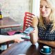 Woman sitting in a cafe with a coffee cup on the table in front of her puts her phone on a payment terminal held by a waitress.