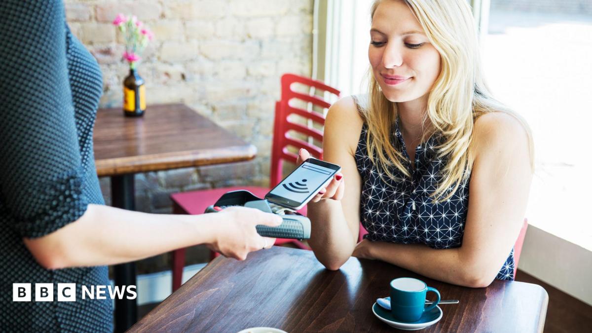 Woman sitting in a cafe with a coffee cup on the table in front of her puts her phone on a payment terminal held by a waitress.