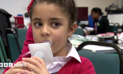 Young child with brown hair, blowing air into plastic measuring device. She wears a red ribbon and red cardigan and a white shirt, She is in school, sitting on a green chair.