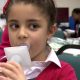 Young child with brown hair, blowing air into plastic measuring device. She wears a red ribbon and red cardigan and a white shirt, She is in school, sitting on a green chair.