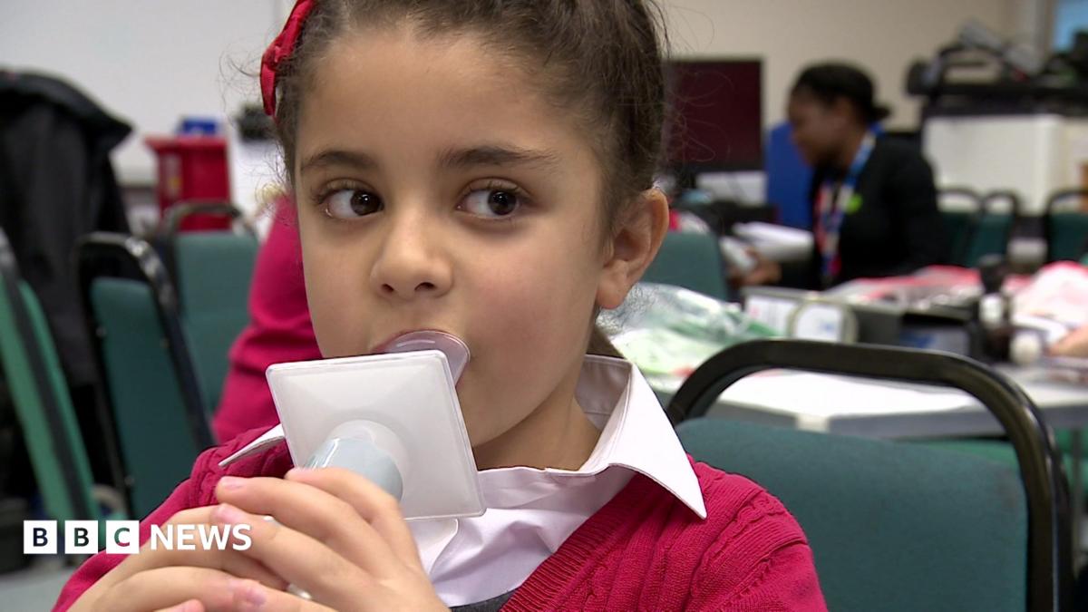 Young child with brown hair, blowing air into plastic measuring device. She wears a red ribbon and red cardigan and a white shirt, She is in school, sitting on a green chair.