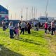 A crowd of people stands on a grassy area beside Clyde Marina, with yachts and sailboat masts visible behind a fence in the background. A large blue building marked “CLYDE MARINA” stands to the left, while people gather along the roadside and waterfront under bright sunshine. A speed limit sign and several parked vehicles are visible near the marina entrance.