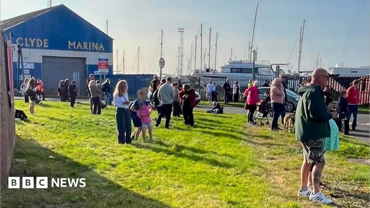 A crowd of people stands on a grassy area beside Clyde Marina, with yachts and sailboat masts visible behind a fence in the background. A large blue building marked “CLYDE MARINA” stands to the left, while people gather along the roadside and waterfront under bright sunshine. A speed limit sign and several parked vehicles are visible near the marina entrance.