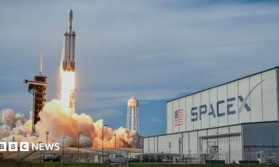 A SpaceX Falcon Heavy rocket blasting off in front of an industrial building with a huge SpaceX logo.