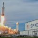 A SpaceX Falcon Heavy rocket blasting off in front of an industrial building with a huge SpaceX logo.
