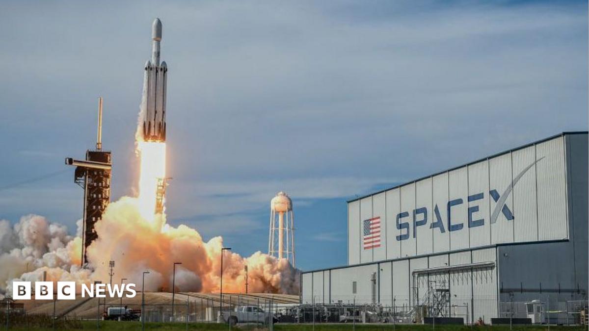 A SpaceX Falcon Heavy rocket blasting off in front of an industrial building with a huge SpaceX logo.