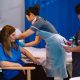 This image shows a healthcare setting with a seated adult receiving an injection in the upper arm from a healthcare worker wearing gloves and a disposable protective apron. A second healthcare worker stands nearby holding paperwork or a clipboard, appearing to document or monitor the procedure. The scene takes place beside a hospital-style bed and is partially enclosed by blue privacy curtains. The color palette is dominated by blue, grey, and white tones, with bright indoor lighting illuminating the room and medical equipment visible in the background. The photograph focuses on the administration of a vaccine and the interaction between healthcare staff and the seated patient.