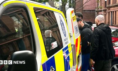 A man is escorted into the back of a blue, white and yellow Police Scotland van by two plain-clothes police officers on a residential street. Cars are parked nearby.