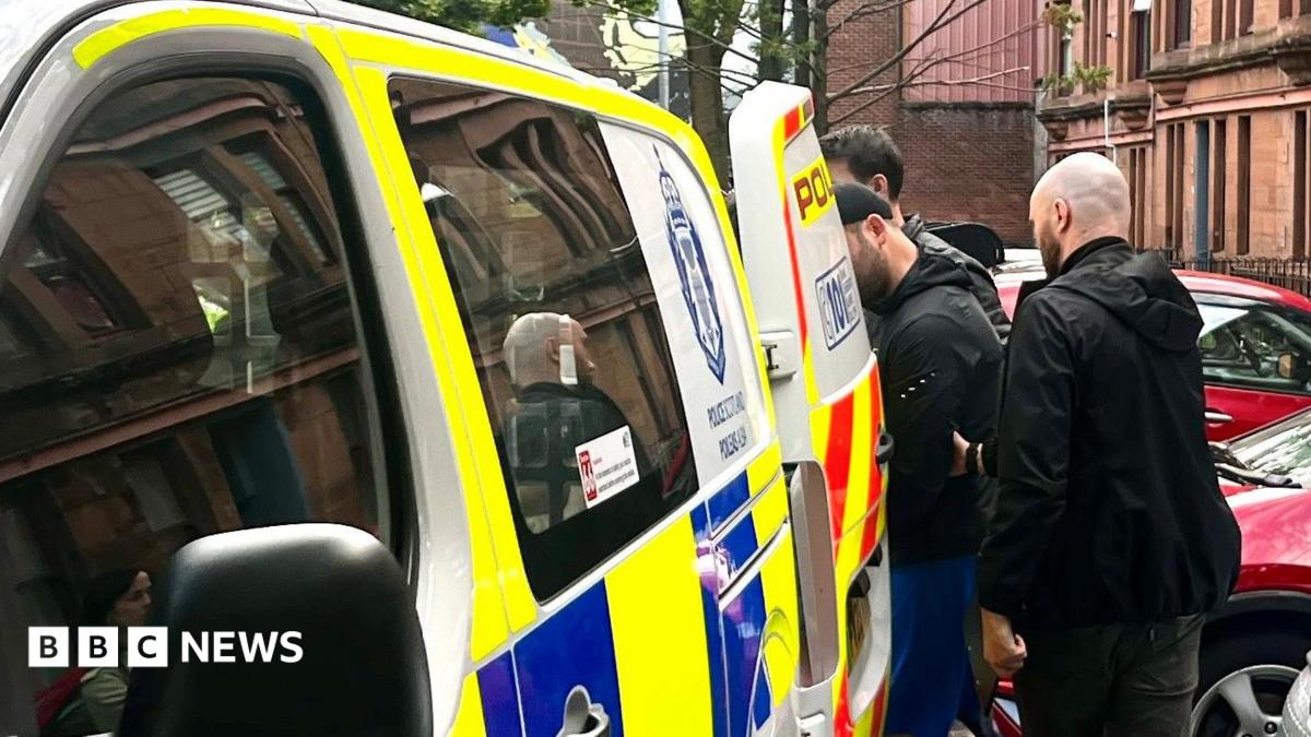 A man is escorted into the back of a blue, white and yellow Police Scotland van by two plain-clothes police officers on a residential street. Cars are parked nearby.