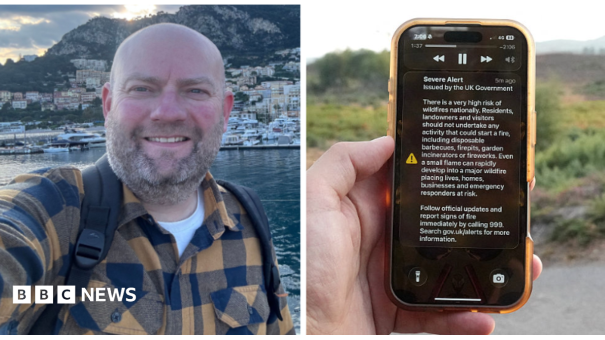 A man holds a phone in front of a green field. The phone is displaying the UK government wildfire alert.