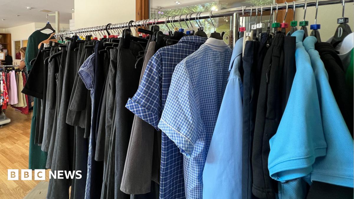 The image shows clothing racks inside a charity shop displaying school uniforms and other clothes, with shirts, skirts and trousers hanging on rails in a bright indoor space.
