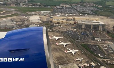 View from a plane shows blue engine of plane and four aircraft on tarmac outside low-rise terminal buildings.