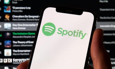 A photo of a hand holding a phone which has the green Spotify logo on it. Behind is a Spotify playlist in the background.