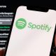 A photo of a hand holding a phone which has the green Spotify logo on it. Behind is a Spotify playlist in the background.