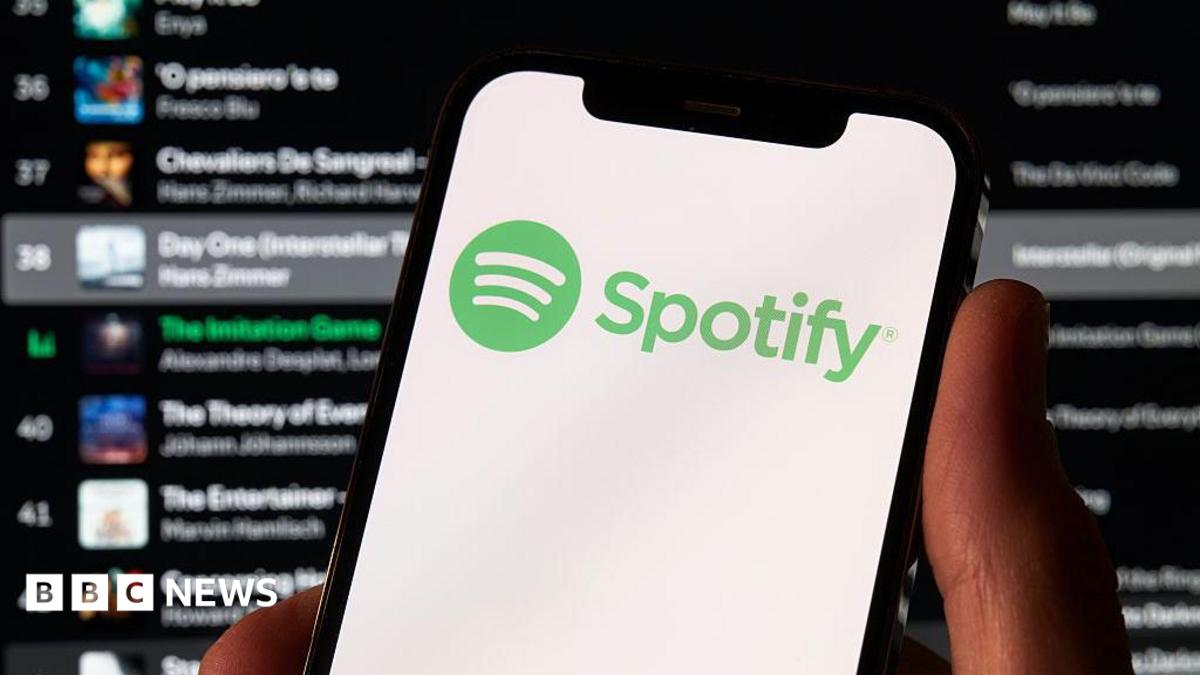 A photo of a hand holding a phone which has the green Spotify logo on it. Behind is a Spotify playlist in the background.