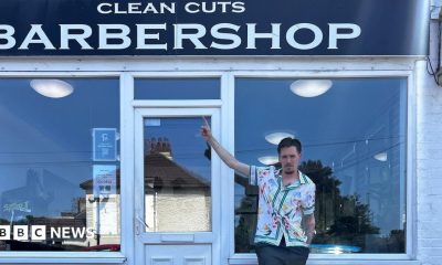 A man in a white floral shirt standing in front of a barber shop. He is pointing at the sign.