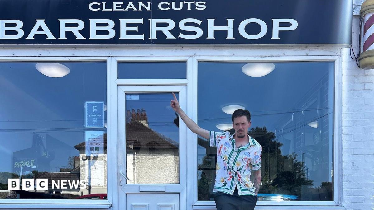A man in a white floral shirt standing in front of a barber shop. He is pointing at the sign.