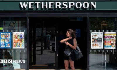 A woman dressed in a black vest and shorts stands outside a Wetherspoons holding her phone.