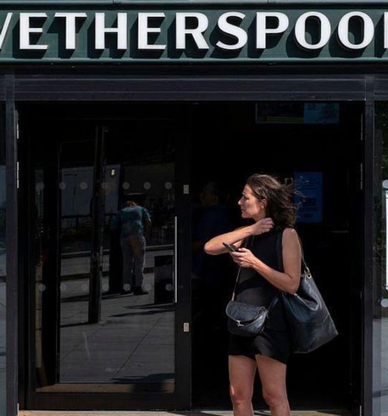 A woman dressed in a black vest and shorts stands outside a Wetherspoons holding her phone.