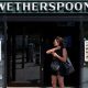 A woman dressed in a black vest and shorts stands outside a Wetherspoons holding her phone.