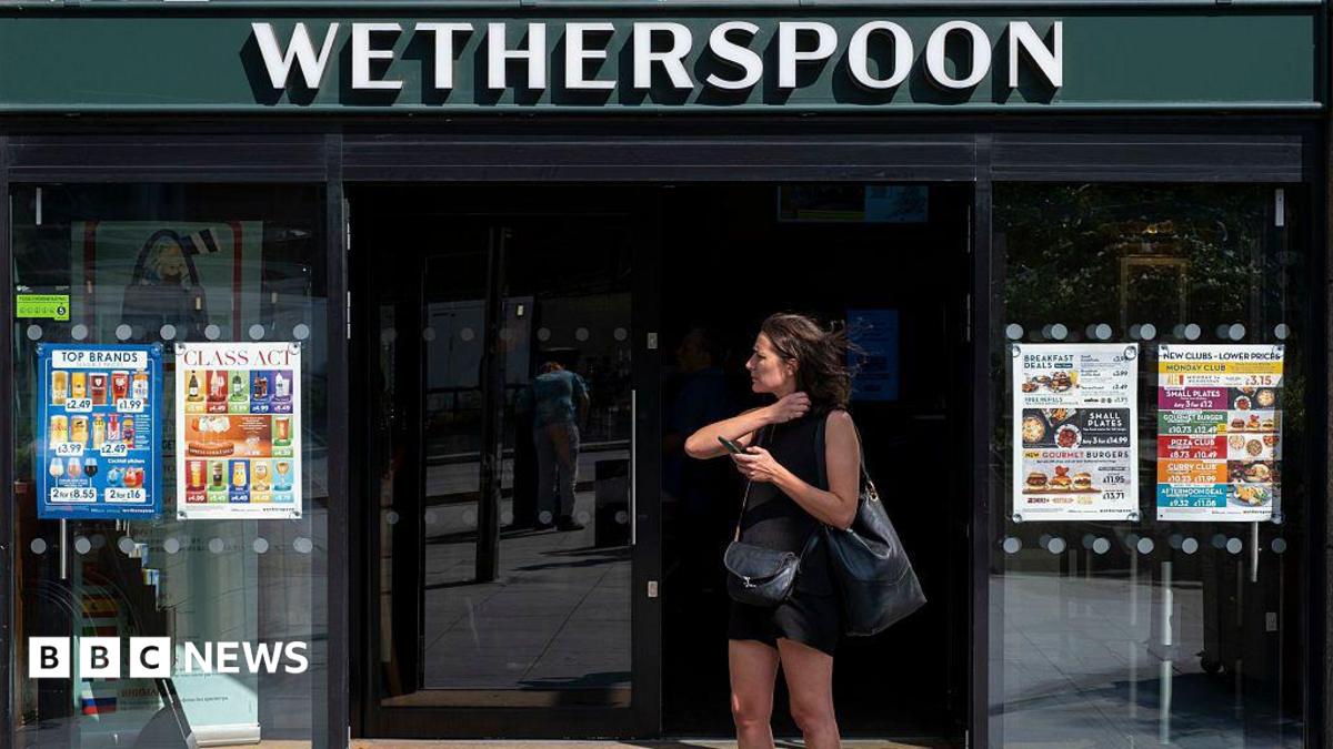 A woman dressed in a black vest and shorts stands outside a Wetherspoons holding her phone.