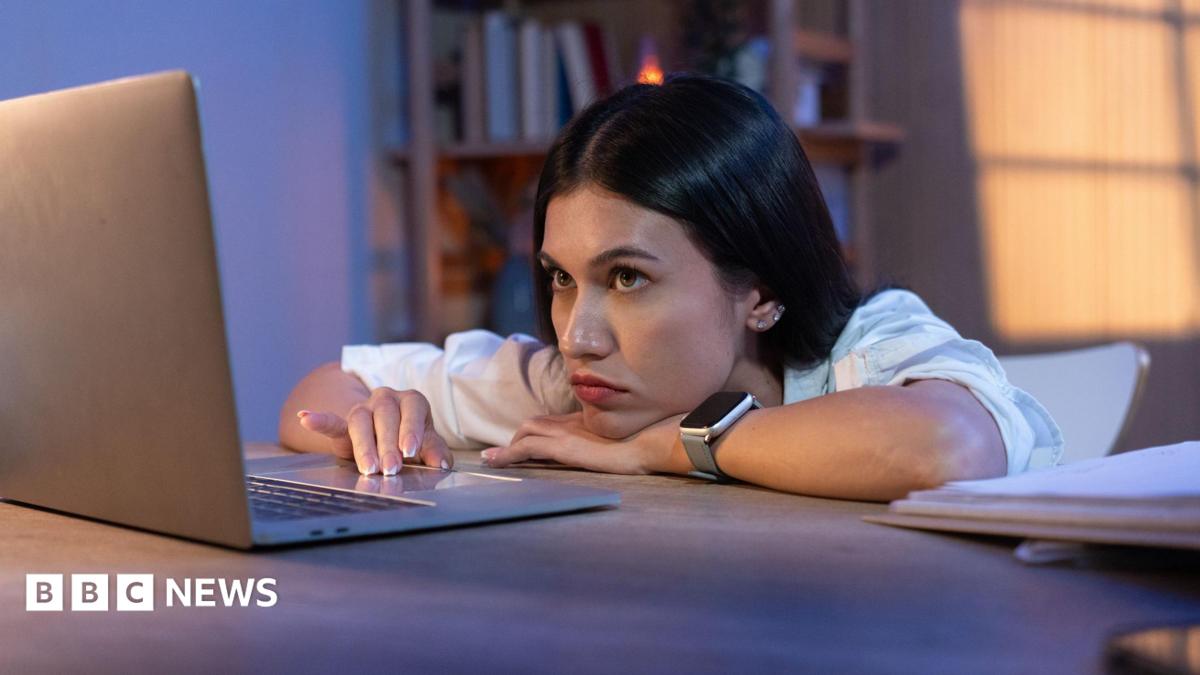 A forlorn young woman slumped over a desk looking at a laptop