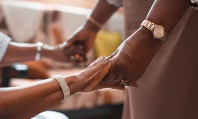Assisted dying: A close-up of two women holding hands, one sat down in a hospital gown, and the other stood up. Both have gold and silver bracelets/ watches on their wrists.