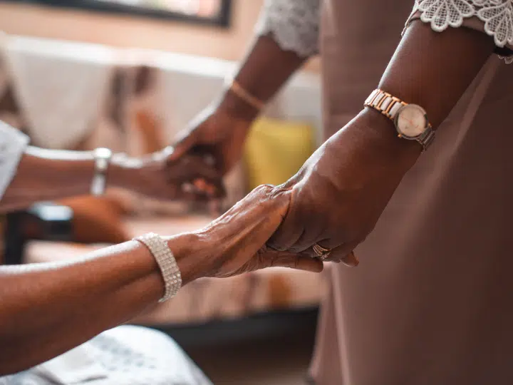 Assisted dying: A close-up of two women holding hands, one sat down in a hospital gown, and the other stood up. Both have gold and silver bracelets/ watches on their wrists.