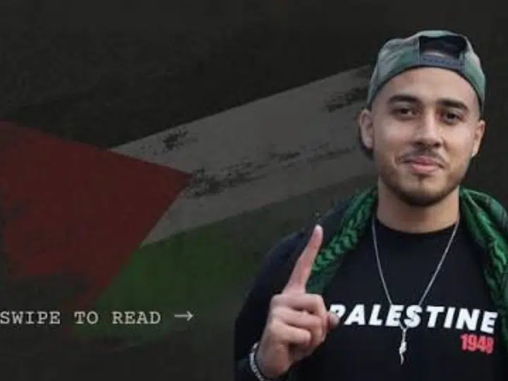 Ahmad Hajahmad Jarrar, a pro-Palestine activist, smiles with his index finger pointing up in a baseball cap turned backwards and a scarf draped on his shoulders. His jumper has 'Palestine' printed on it