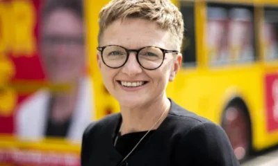 New Greater Manchester mayor, Bev Craig, smiling with her signature cropped hair and round-rimmed glasses in front of a campaign bus
