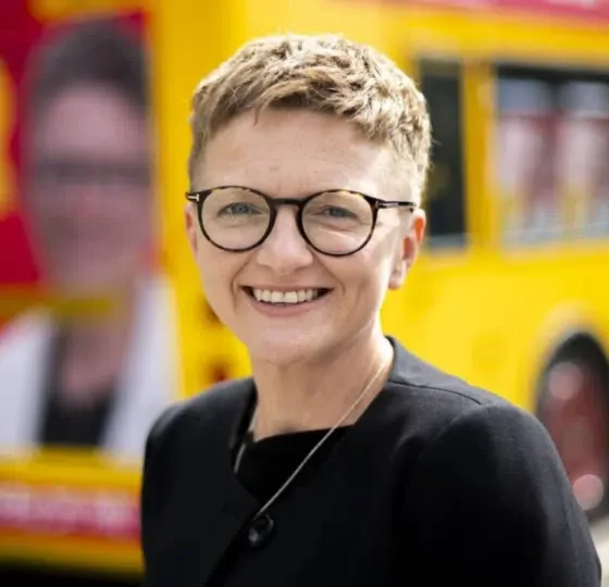 New Greater Manchester mayor, Bev Craig, smiling with her signature cropped hair and round-rimmed glasses in front of a campaign bus