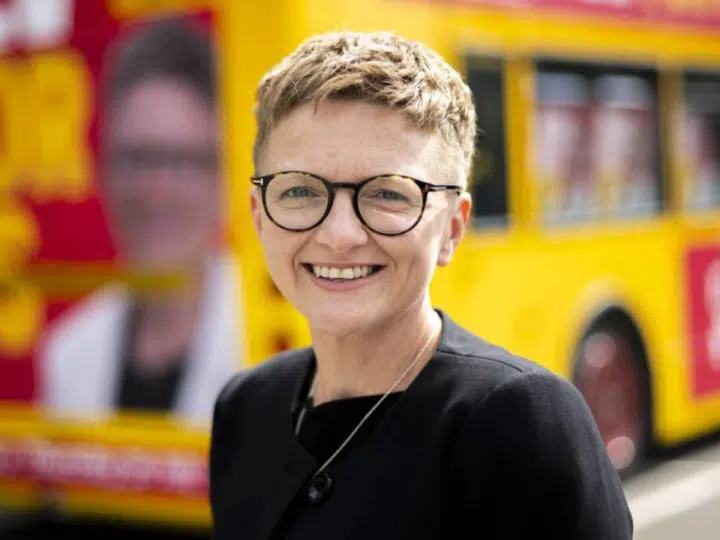New Greater Manchester mayor, Bev Craig, smiling with her signature cropped hair and round-rimmed glasses in front of a campaign bus