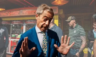Nigel Farage, leader of Reform UK, reacts to a question from journalists in Clacton and he's captured with both hands up, palms facing outwards. An amusement park games machine is behind him.