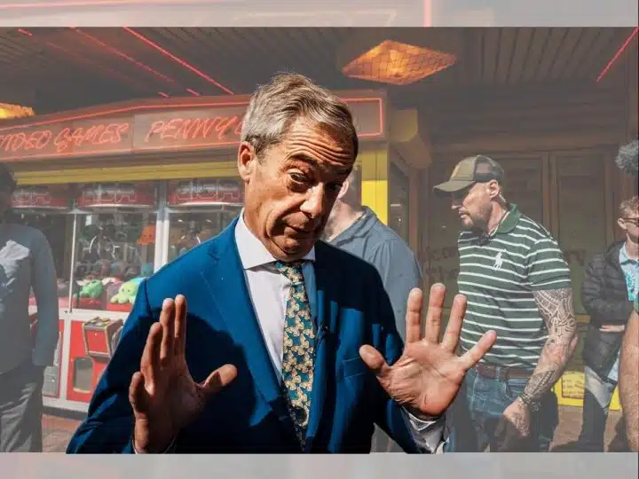 Nigel Farage, leader of Reform UK, reacts to a question from journalists in Clacton and he's captured with both hands up, palms facing outwards. An amusement park games machine is behind him.