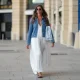 Amanda Derhy wears earrings, sunglasses, a blue denim jacket, a white summer maxi pleated low-neck slit off-shoulder dress, sandals, during a street style fashion photo session, on August 20, 2022 in Paris, France.