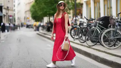 Maria Rosaria Rizzo wears long straight brown hair partially covered with a beige Borsalino hat, oversized black sunglasses, a red Mango sleeveless midi dress, a red Moschino sweater worn over the shoulders, a Chloe straw bag with white leather straps, and white Lacoste tennis sneakers shoes, during a street style fashion photo session, on April 18, 2026 in Paris, France.
