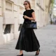 Diane Batoukina wears long straight brown hair, black sunglasses by Kaleos, a black knit sleeveless sweater by St. Agni, a black leather midi skirt by The Frankie Shop, a black top-handle bag Kelly by Hermes, black pointed shoes by COS, during a street style fashion photo session, on April 13, 2026 in Paris, France.