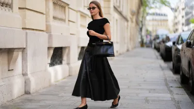 Diane Batoukina wears long straight brown hair, black sunglasses by Kaleos, a black knit sleeveless sweater by St. Agni, a black leather midi skirt by The Frankie Shop, a black top-handle bag Kelly by Hermes, black pointed shoes by COS, during a street style fashion photo session, on April 13, 2026 in Paris, France.
