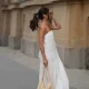 Aylin Sengül is seen wearing a white spaghetti-strap maxi dress with textured horizontal panels and a gathered hem by COS; brown leather “Oran” sandals by Hermès; a raffia shoulder bag with a slouchy hobo shape by COS; a delicate gold-tone necklace; a rose gold “Knot” bracelet by Celine; a vintage tennis bracelet; a slim silver-tone bangle; a gold ring; slim oval dark tortoiseshell sunglasses by Chanel during Berlin Fashion Week Spring/Summer 2027 on July 4, 2026 in Berlin, Germany.