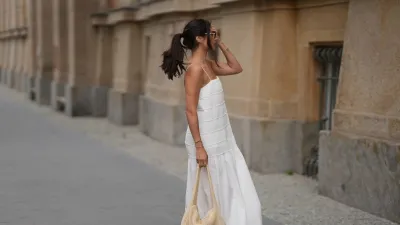 Aylin Sengül is seen wearing a white spaghetti-strap maxi dress with textured horizontal panels and a gathered hem by COS; brown leather “Oran” sandals by Hermès; a raffia shoulder bag with a slouchy hobo shape by COS; a delicate gold-tone necklace; a rose gold “Knot” bracelet by Celine; a vintage tennis bracelet; a slim silver-tone bangle; a gold ring; slim oval dark tortoiseshell sunglasses by Chanel during Berlin Fashion Week Spring/Summer 2027 on July 4, 2026 in Berlin, Germany.