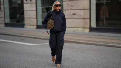 Mirja Klein is seen wearing a navy zip front jacket with a high collar from The Frankie Shop; matching navy cargo trousers with a self tie belt from The Frankie Shop; brown leather flip flops from A.Emery; a medium sized olive suede top handle bag from DeMellier; oversized rectangular acetate sunglasses with dark tinted lenses from Phoebe Philo; blonde hair worn long with a center part during the Copenhagen Fashion Week Spring/Summer 2027 on August 6, 2026 in Copenhagen, Denmark.