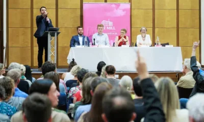 From behind the rows of people, the Manchester politicians are photographed on a long table with mics on the stage while a man, seemingly the host, points at the crowd with their hands up at Mill Media's hustings