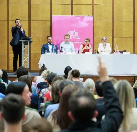 From behind the rows of people, the Manchester politicians are photographed on a long table with mics on the stage while a man, seemingly the host, points at the crowd with their hands up at Mill Media's hustings