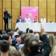 From behind the rows of people, the Manchester politicians are photographed on a long table with mics on the stage while a man, seemingly the host, points at the crowd with their hands up at Mill Media's hustings