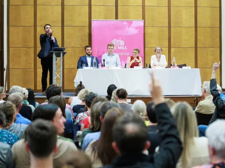 From behind the rows of people, the Manchester politicians are photographed on a long table with mics on the stage while a man, seemingly the host, points at the crowd with their hands up at Mill Media's hustings