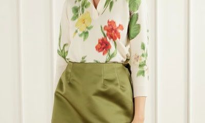 A woman wearing a floral top (white, red, green, and yellow) and olive green skirt